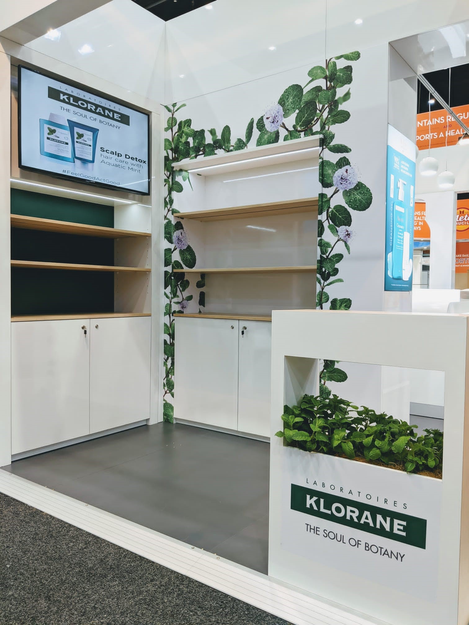 Klorane stand with television turned on showing feature 'Scalp Detox' products and shelves surrounded by green vinery wallpaper with white flowers